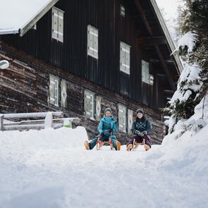 Zwei Frauen sitzen auf ihren Rodeln und starten auf der Rodelbahn. Im Hintergrund ist ein altes Holzgebäude zu sehen. | © TVB Wipptal | Johannes Bitter