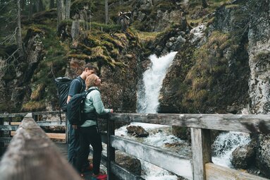 Eine Frau und ein Mann stehen auf einer Holzbrücke und schaut zum Wasserfall hinauf | © TVB Wipptal | Johannes Bitter