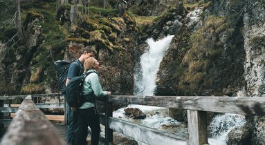 Eine Frau und ein Mann stehen auf einer Holzbrücke und schaut zum Wasserfall hinauf | © TVB Wipptal | Johannes Bitter