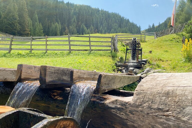 Eine Wassermühle aus Holz treibt Wasser in zwei Holzträge, die in einen Brunnen münden | © Daniela Plattner