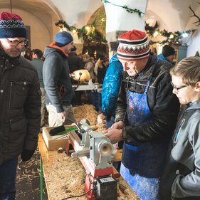 Ein Mann drechselt Holz und ein Mann und ein Junge schauen zu. | © Kreativstadl