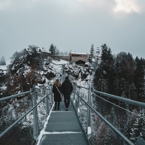 Ein Mann und eine Frau gehen nebeneinander in verschneiter Landschaft auf einer Hängebrücke auf ein  | © Helena Beermeister