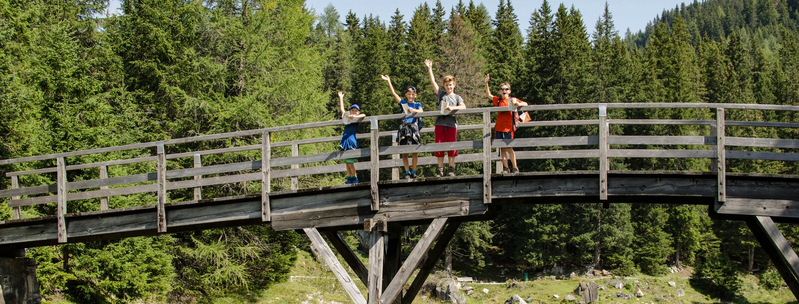 Vier Kinder winken von einer Holzbrücke, die über einen See im Wald gespannt ist | © TVB Wipptal | Joakim Strickner