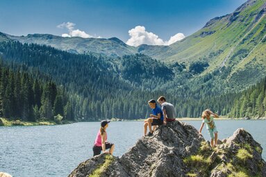 Eine Frau und drei Kinder kraxeln auf einen kleinen Felsen vor einem Bergsee | © TVB Wipptal | Joakim Strickner