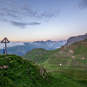 Frau steht bei einem Gipfelkreuz kurz vor Sonnenaufgang mit Blick auf die umliegenden Berge | © TVB Wipptal | Judith Hammer